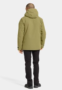Veste imperméable vert olive avec capuche, texture douce, manches longues, poignets élastiques et un logo bien visible dans le dos. Pantalons et bottes noirs.