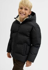 Smilende blond dreng iført en sort puffjakke med hætte og brune bukser, stående imod en ensfarvet hvid baggrund.
