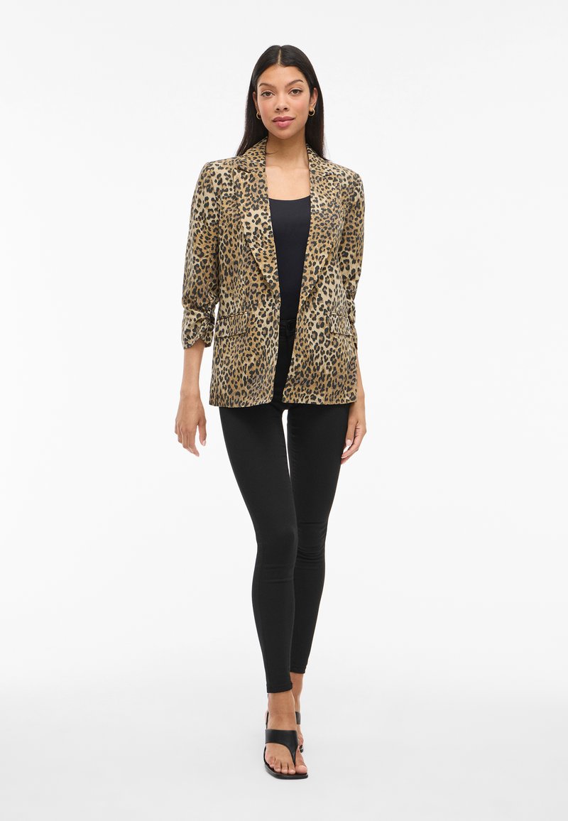 Leopardenmuster Blazer aus strukturiertem Stoff, mit einem schmalen Kragen, zwei Vorderaschen und hochgerollten Ärmeln. Getragen über einem schwarzen Top und Leggings.
