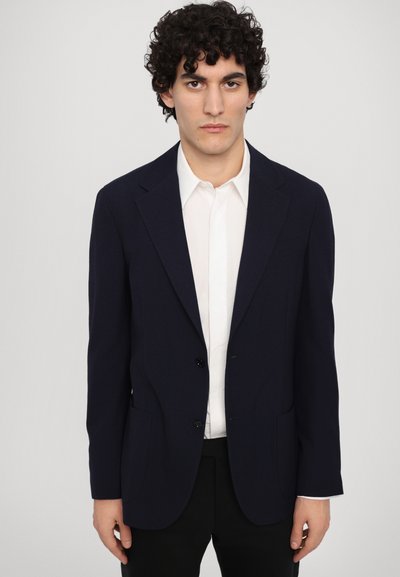 Blazer blu navy con due bottoni, rever a punta e tasche a toppa. Indossato su una camicia bianca, presenta una texture liscia e una vestibilità su misura.