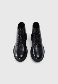 Botas de cuero negras hasta el tobillo con puntera redonda, que presentan una textura suave, seis ojales metálicos y cordones negros. Suelas de goma para mayor tracción.