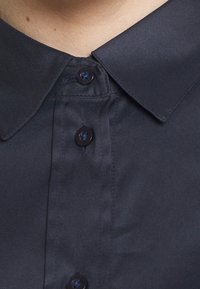 Marinblå skjorta med spetsig krage, prydd med svarta knappar och en slät satintextur. Har en lätt glans och rena sömmar.