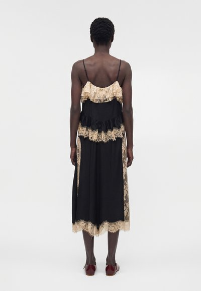 Άτομο που στέκεται με πλάτη στην κάμερα, φορώντας μαύρο μίντι φόρεμα με μπεζ δαντέλα και κόκκινα φλατ παπούτσια σε μονόχρωμο φόντο.