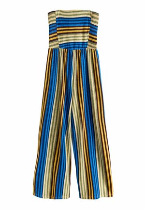 Kobieta stoi oparta o ścianę, ubrana w bezramienny kombinezon z szerokimi nogawkami w pionowe paski w kolorach niebieskim, żółtym i brązowym oraz złote sandały.