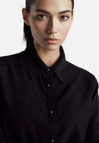Czarna koszula z kołnierzykiem, zapinana na guziki z przodu, ozdobiona drobnymi srebrnymi akcentami. Gładka faktura, prosty fason.