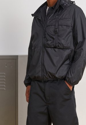 Mężczyzna w czarnej kurtce zapinanej na zamek błyskawiczny z kieszeniami na piersi i bokach, stojący z jedną ręką w kieszeni czarnych spodni na jasnym tle.