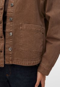 Καφέ μπουφάν από τζιν με κλείσιμο με κουμπιά, μια μεγάλη μπροστινή τσέπη και άνετη γραμμή. Το ύφασμα έχει ελαφρώς υφασμένη υφή.