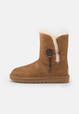 Bruine suède winterlaars met zachte witte fleece voering, ronde neus, platte zool, en decoratieve knoop met bijgevoegde hanger aan de zijkant.