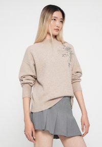 Maglione beige a maglia con un collo alto, caratterizzato da decorazioni argentate su una spalla, abbinato a una gonna grigia plissettata.