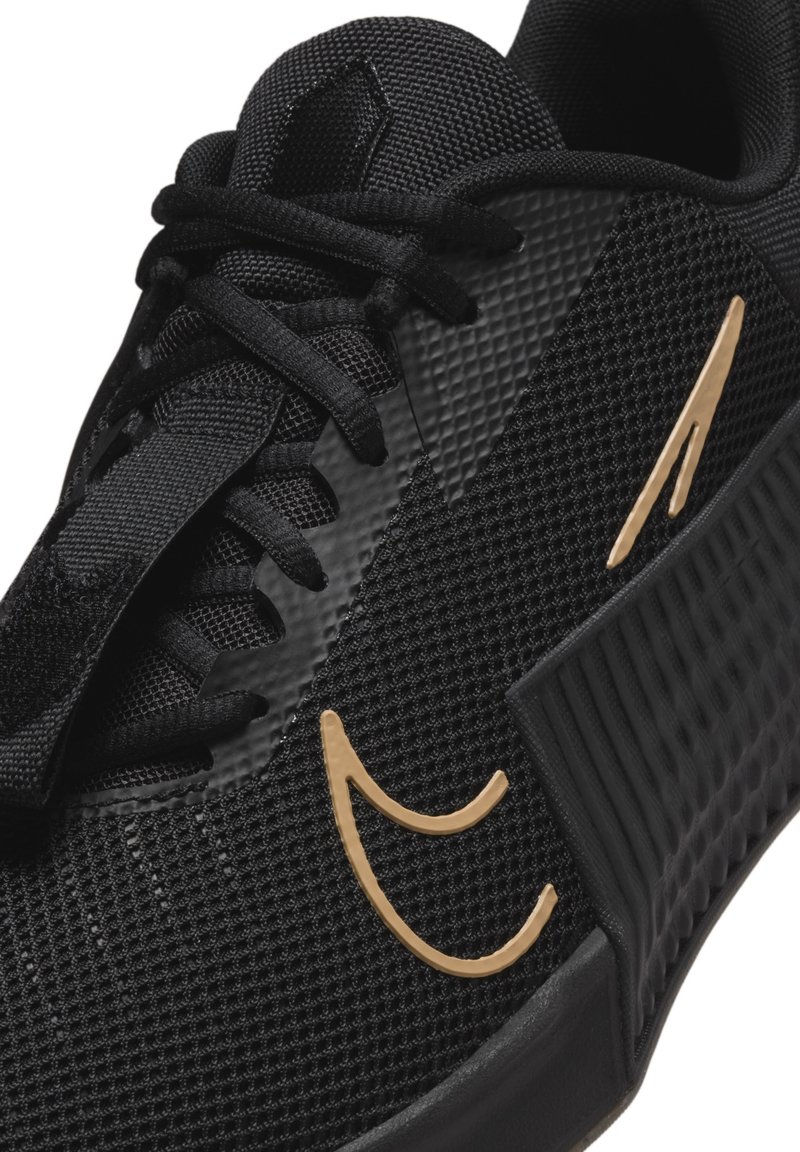 Zwarte sportieve schoen met een gestructureerde mesh bovenkant, platte zwarte veters en een opvallend gouden logo aan de zijkant. Beschikt over een ribbelige zwarte zool.