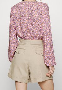 Blusa floreale a maniche lunghe con motivi rosa, arancioni e verdi; abbinata a pantaloni corti crema con due tasche posteriori e vita con cintura.