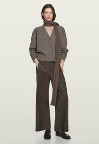 Hnědý vlněný kardigan s výstřihem do V a zapínáním na knoflíky, spojený se širokými hnědými kalhotami. Doplňuje ho žebrovaný hnědý šátek. Černé boty.