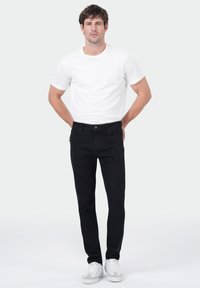 Homme se tenant contre un fond blanc, portant un t-shirt blanc, un jean noir et des baskets blanches, les mains derrière le dos.