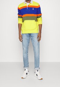 Pullover colorato con righe orizzontali in blu, arancione, verde e giallo, con tasca frontale e colletto sopra una camicia bianca. Indossato con jeans in denim chiaro e sneaker bianche.