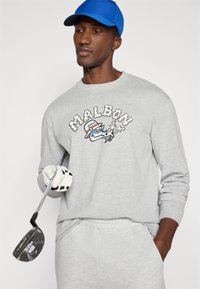 Γκρίζα μακρυμάνικη μπλούζα με γραφικό "MALBON", φορεμένη με μπλε καπέλο. Κρατάει ένα γκολφ κλαμπ, ο γαντωμένος του χέρι είναι ορατό. Χαλαρή αθλητική ένδυση.