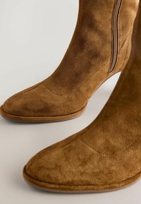 Bruna mockaskinnsankelstövlar med spetsig tå, sidodragkedja och slät yta. Har detaljer med sömmar och en lätt klack.