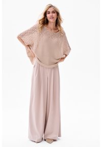Beige gebreide top met een gestructureerde, open weefstijl en brede mouwen, gecombineerd met luchtige, lichtroze wijde broek en neutrale schoenen.