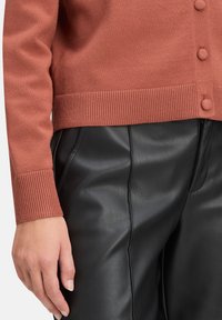 Rdzawy kardigan z prążkowanym dołem i mankietami, z okrągłymi guzikami. Połączony z błyszczącymi czarnymi spodniami z ekoskóry.