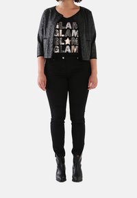 A woman stands in stylish black clothing, featuring a glittery "GLAM" printed top, black jacket, jeans, and ankle boots, against a white background.