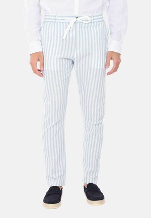 Uomo che indossa pantaloni a righe verticali azzurro chiaro e bianchi con coulisse bianca, camicia bianca e scarpe slip-on blu scuro.