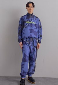 Mėlynas treniruočių kostiumas su lapų raštais, kuriame yra zip-up džemperis ir elastingu juosmeniu kelnės, dekoruotos baltomis detalėmis. Juodi slip-on batai.