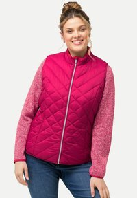 Fuchsia gesteppte Weste mit Stehkragen und Frontreißverschluss, kombiniert mit rosa strukturierten Ärmeln. Getragen über blauen Jeans.