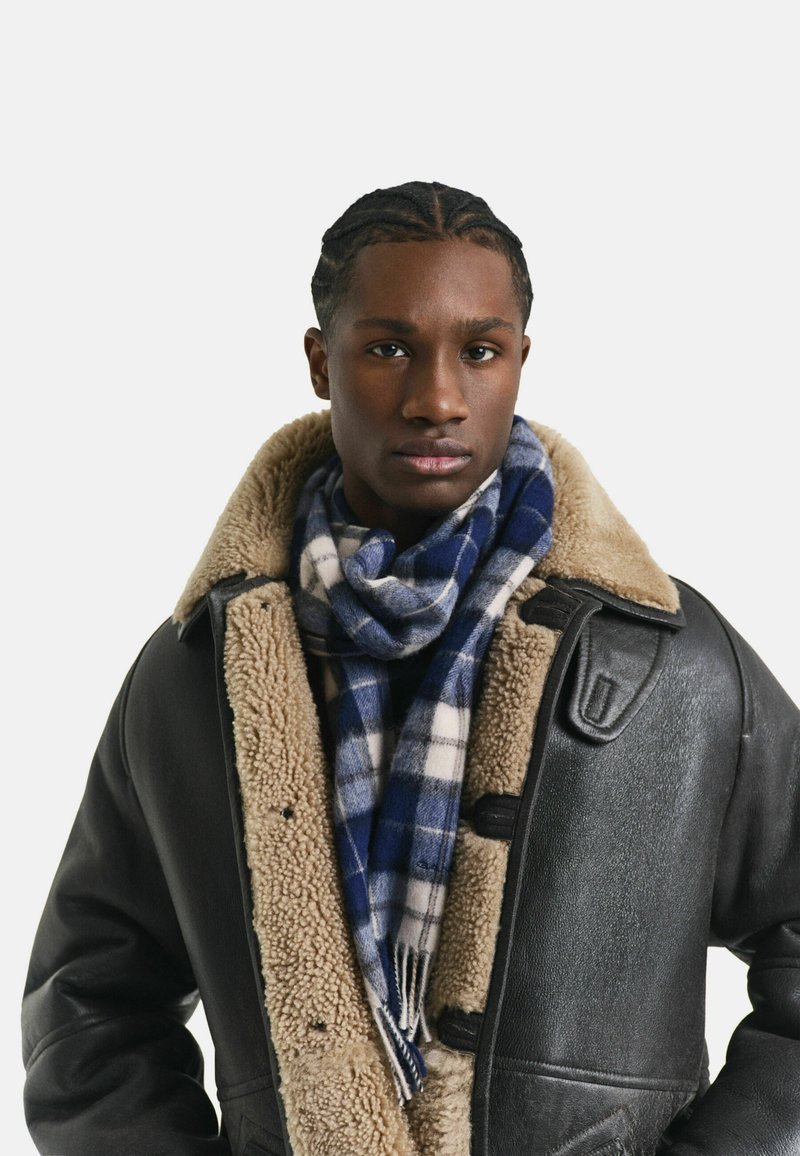 Jeune homme avec des cheveux tressés portant une veste en cuir noir avec un intérieur en laine beige et une écharpe à carreaux bleus et blancs.
