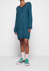 Abito in maglia verde acqua con scollatura a V, maniche lunghe e orlo a costine. Abbinato a sneakers multicolori e calzini a manica corta rosa.