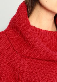 Rode gebreide trui met een grote, gestructureerde col. De stof vertoont een ribbelpatroon, dat warmte en zachtheid benadrukt.