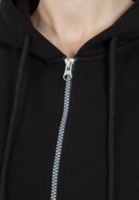 Il cappuccio zip-up nero presenta una cerniera argentata, lacci intrecciati e una texture di tessuto morbido. Il design include un cappuccio e un collo aderente.