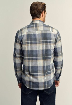 Man wearing blue, beige, and white plaid long-sleeve shirt and dark jeans, standing facing away against a plain background.