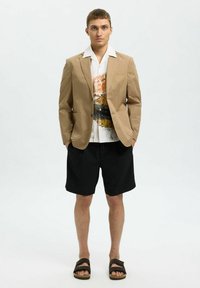 Světlý hnědý blazer přes bílo-oranžovou vzorovanou košili, spárovaný s černými šortkami a hnědými sandály, umístěný na jednobarevném pozadí.