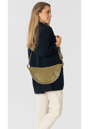 Olivengrüne Leder-Schultertasche mit geschwungenem Design, silbernem Ankeranhänger und verstellbarem Tragegurt, getragen zu einem dunklen Outfit und hellen Hosen.