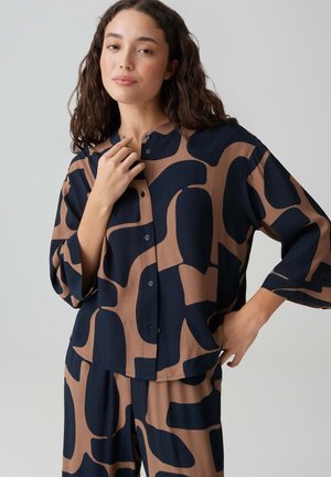 Frau mit dunklem lockigem Haar, die ein passendes, locker sitzendes Hemd und Hose mit abstraktem schwarz-braunem Muster trägt und vor einem einfarbigen Hintergrund posiert.