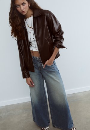 Giovane donna con lunghi capelli, che indossa una giacca di pelle marrone scuro, un top corto bianco, jeans a gamba larga sbiaditi blu, collana e sneaker.