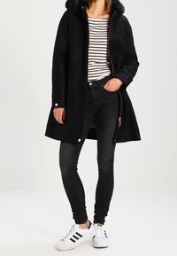 Donna che indossa un cappotto cappuccio nero, una maglietta a righe, jeans skinny neri e sneakers bianche Adidas con strisce nere, in piedi con una mano in tasca.