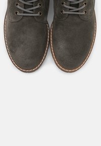 Zapatos de ante gris con punteras redondas, cordones grises y ojales de metal. La suela presenta un borde cosido en un color contrastante.