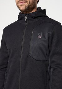 Zwarte fleece jack met een getextureerde oppervlakte, ritssluiting op de borstzak en capuchon. Bevat het spinnenlogo op de linkerbovenkant.