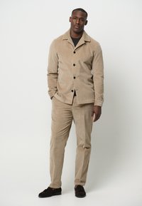 Beige corduroy jas met kraag en zes knopen, gecombineerd met bijpassende corduroy broek. Beide hebben een verticale ribbelstructuur.