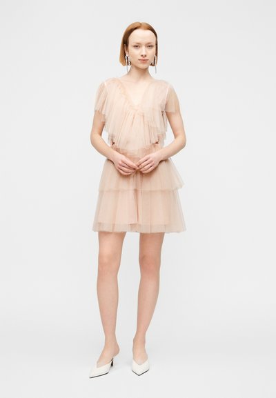 Γυναίκα με στρωματωτό φόρεμα από γυμνό τούλι με κοντά μανίκια, όρθια με τα χέρια σταυρωμένα, φορώντας εντυπωσιακά σκουλαρίκια και λευκά μυτερά τακούνια.