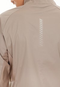 Ljusbeige jacka med en slät yta, som har ett vertikalt reflexdesignelement på baksidan och kontrasterande ljusbruna ärmar.