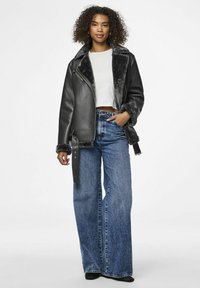 Giacca di pelle nera con colletto in pelliccia sintetica, abbinata a jeans blu ampi a vita alta. La modella è in piedi contro uno sfondo chiaro.