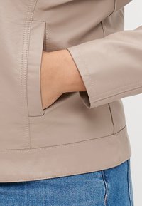 Χέρι μέσα στη πλαϊνή τσέπη μιας μπεζ δερμάτινης ζακέτας που φοριέται πάνω από μπλε τζιν παντελόνι.