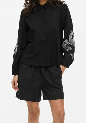 Person iført sort langærmet skjorte med hvidt blomsterbroderi på ærmerne og sorte shorts, stående med den ene hånd i lommen.