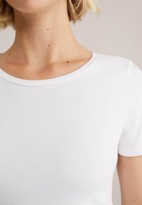 Nahaufnahme eines Halses und oberen Brustbereichs einer Person, die ein schlichtes weißes Kurzarmhemd trägt, vor einem neutralen Hintergrund.