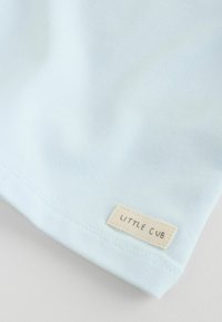 Tissu de coton bleu clair à la texture lisse, avec une petite étiquette tissée portant l'inscription "LITTLE CUB" cousue sur le bord inférieur.