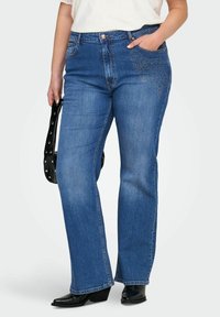 Žena v modrých bootcut džínách s květinovými kamínkovými detaily na kapsách, bílém topu, černých botách a držící černou kabelku s hroty.