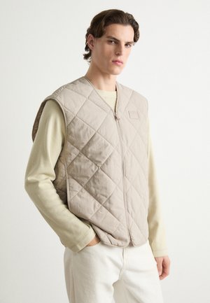 Lichtbeige gewatteerde vest met een rits aan de voorkant en een subtiele logo patch, gedragen over een lichtgroen overhemd met lange mouwen, gecombineerd met witte broeken.