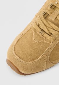Tan suede sneaker featuring a textured upper, reinforced stitching, and a rubber sole. Laces in a matching color with dark stripes.