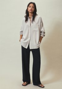 Chemise boutonnée gris clair avec poche, coupe décontractée et manches retroussées, associée à un pantalon large noir et des sandales plates.